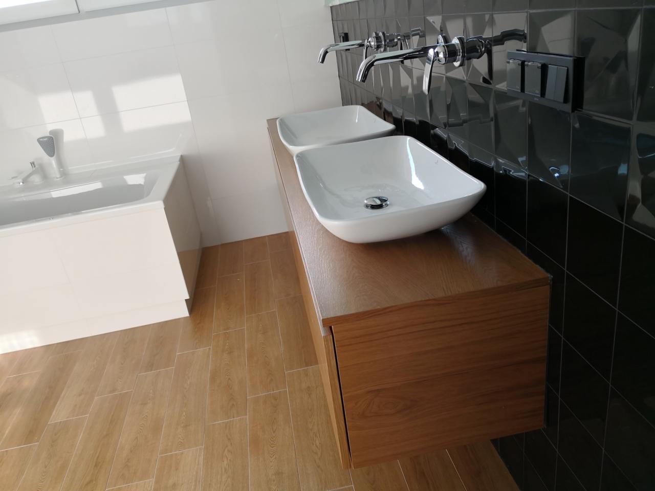 Nowoczesna łazienka z dwiema białymi umywalkami nablatowymi na drewnianej szafce, czarne płytki na ścianie i jasne płytki podłogowe imitujące drewno, bateria umywalkowa w chromie.