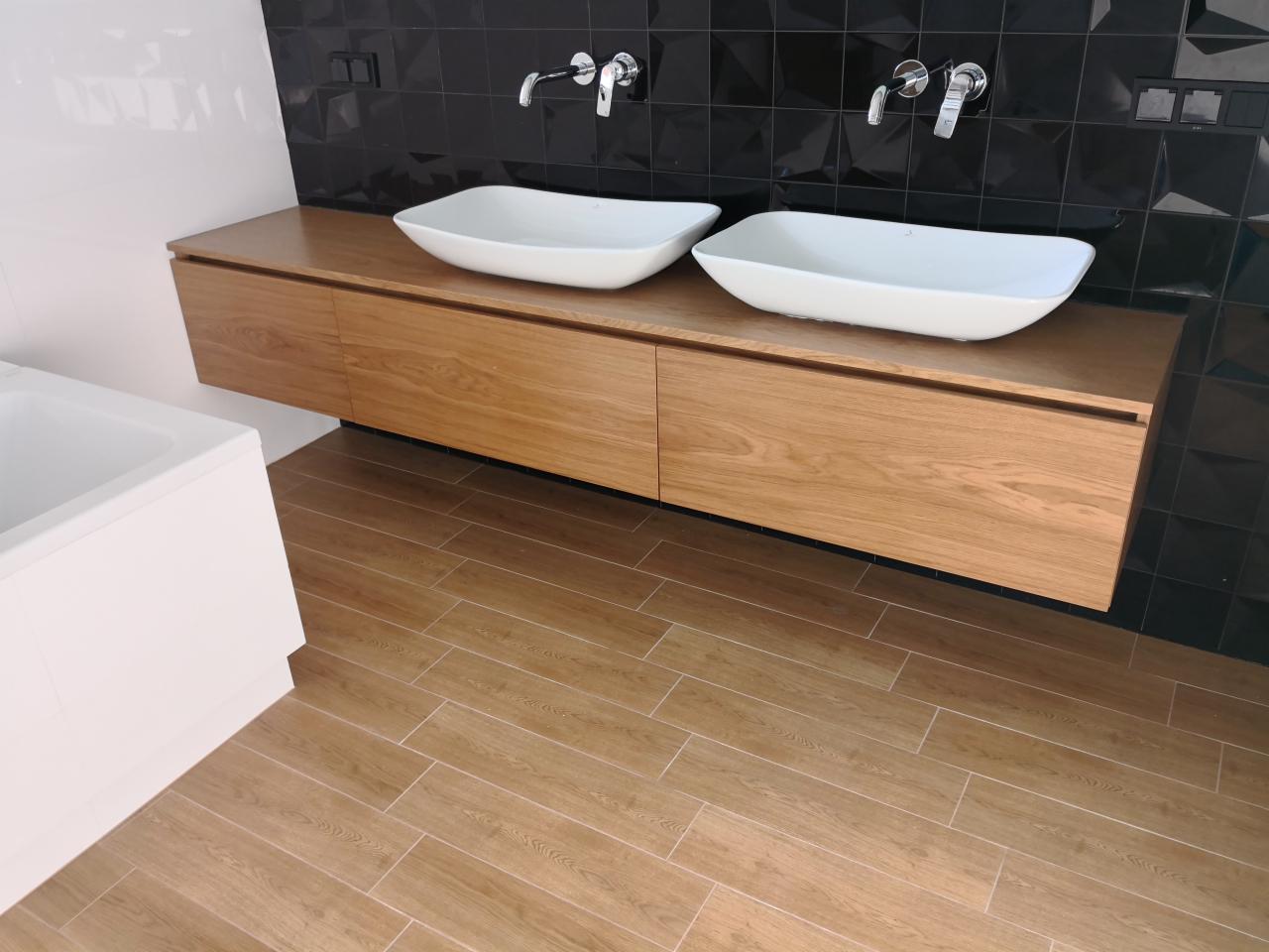 Nowoczesna łazienka z drewnianą szafką pod umywalki nablatowe, czarną mozaiką na ścianie i podłogą wyłożoną płytkami imitującymi drewno.