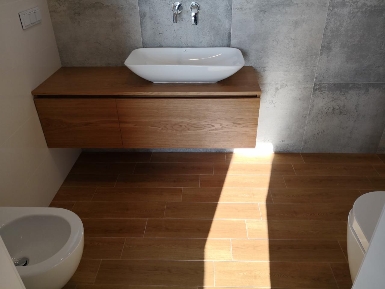 Nowoczesna łazienka z drewnianą szafką pod umywalką, białą umywalką nablatową i szarą ścianą z płytek imitujących beton, uzupełniona podłogą z płytek drewnopodobnych, oświetlona naturalnym światłem...