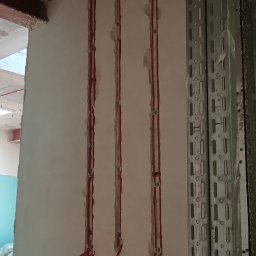 comlive.pl - Czerwone przewody w bruzdach ściany, zabezpieczone metalowymi uchwytami, prowadzone od sufitu. Obok metalowa listwa montażowa z otworami.