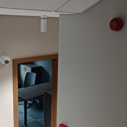 comlive.pl - Wnętrze biura z zainstalowanym systemem alarmowym: czujka ruchu, sygnalizator akustyczny i kamera monitoringu. Widoczny fragment biura z biurkiem i krzesłem.