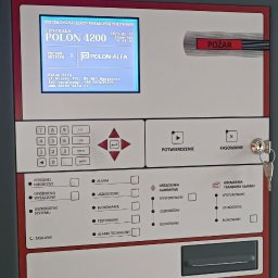 comlive.pl - Centrala systemu sygnalizacji pożarowej Polon 4200 z wyświetlaczem, klawiaturą i przyciskiem 'Pożar'. Widoczne diody statusu i kluczyk w zamku.