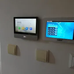 Dwa panele sterowania systemem zabezpieczeń zamontowane na ścianie, jeden z dotykowym ekranem pokazującym menu główne, drugi z ekranem wyświetlającym konfigurację systemu Satel Integra 128-WRL...