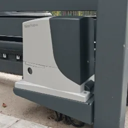 Szara automatyka do bram przesuwnych Nice Robus zamontowana na szynach, widoczna z bliska na tle kostki brukowej.