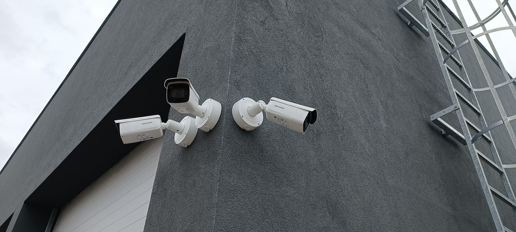 Trzy kamery monitoringu Hikvision zamontowane na szarej, teksturowanej ścianie budynku, widoczne z dołu pod kątem, z metalową drabinką po prawej stronie.