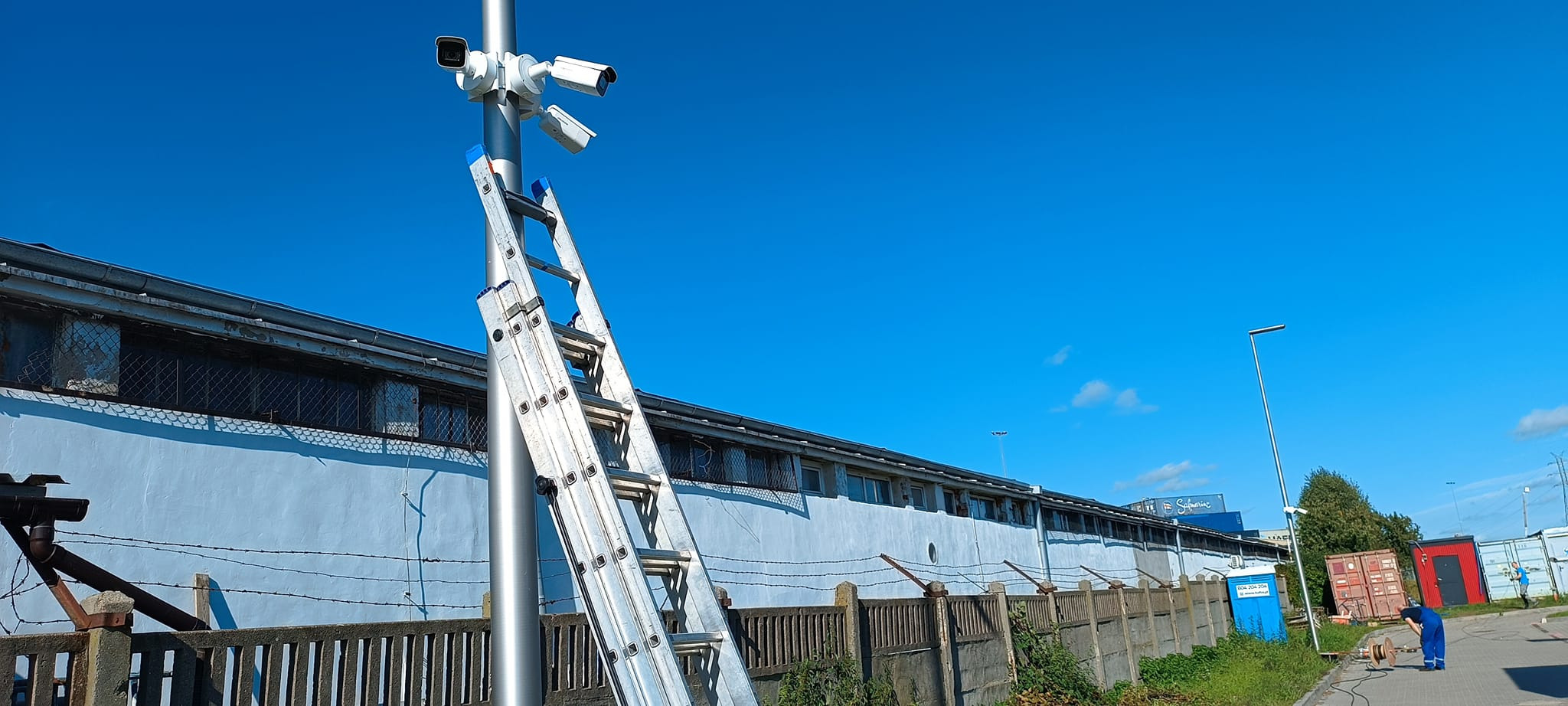 Instalacja czterech kamer monitoringu na metalowym słupie, drabina oparta o słup, budynek w tle z drutem kolczastym na murze.