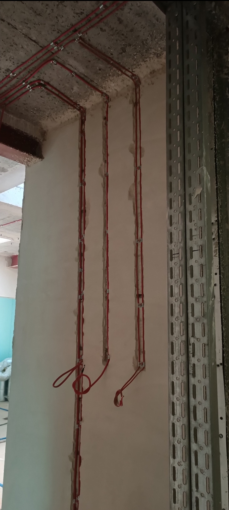 Czerwone przewody w bruzdach ściany, zabezpieczone metalowymi uchwytami, prowadzone od sufitu. Obok metalowa listwa montażowa z otworami.