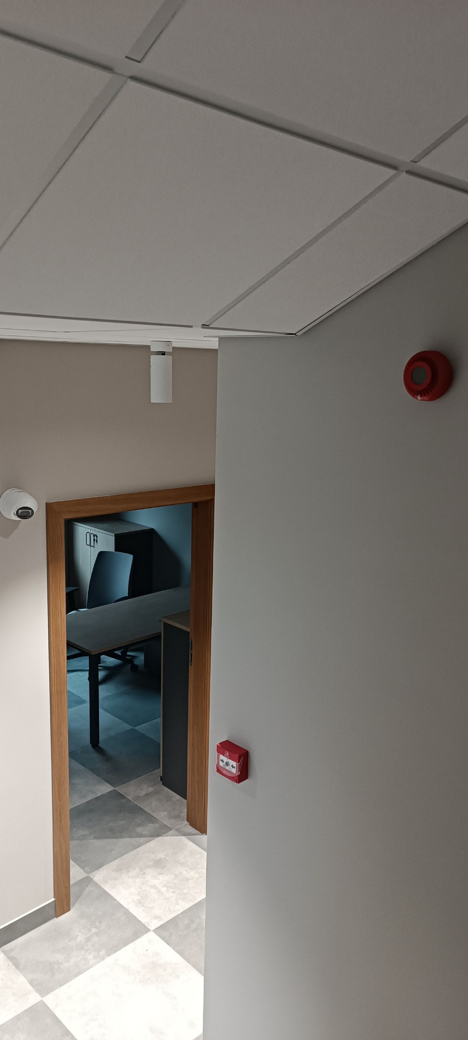 Wnętrze biura z zainstalowanym systemem alarmowym: czujka ruchu, sygnalizator akustyczny i kamera monitoringu. Widoczny fragment biura z biurkiem i krzesłem.