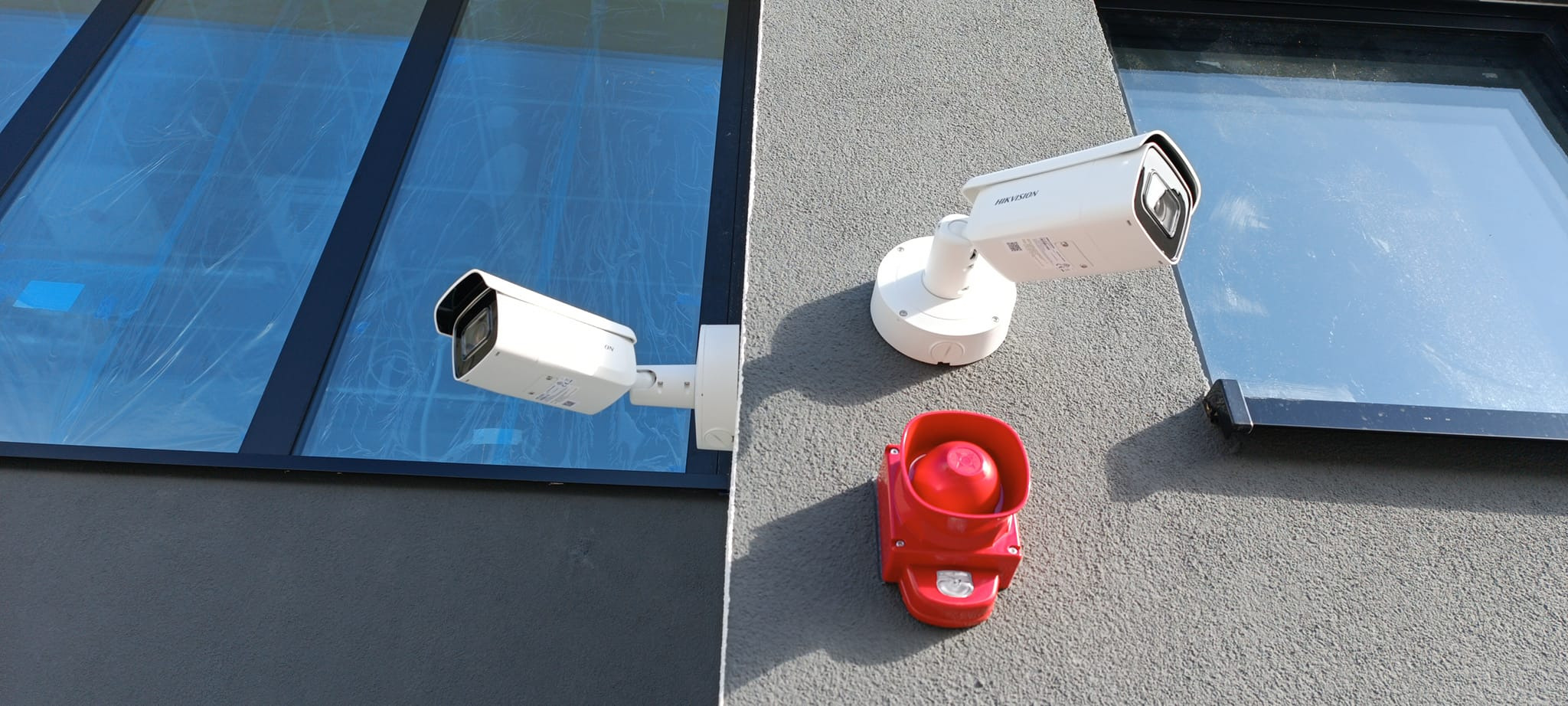 Dwie kamery monitoringu Hikvision i czerwona syrena alarmowa zamontowane na elewacji budynku z oknami. System zabezpieczeń w trakcie instalacji lub po niej.