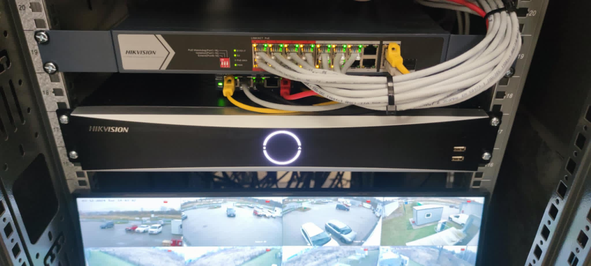 Zamontowany w szafie serwerowej rejestrator Hikvision z podglądem z kamer monitoringu oraz switch sieciowy z podłączonymi kablami.