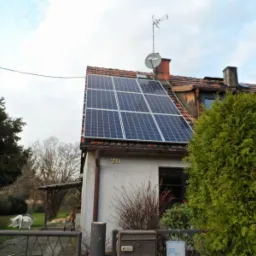 Panele słoneczne na dachu domu jednorodzinnego z anteną satelitarną i telewizyjną, widok z poziomu ulicy w pochmurny dzień.