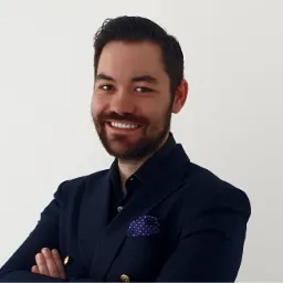 Uśmiechnięty mężczyzna z brodą, ubrany w granatową marynarkę z ozdobnymi guzikami i poszetką w kropki, pozuje z założonymi rękami na jasnym tle.