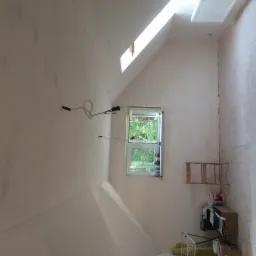 Poddasze w trakcie ocieplania: widoczne okno dachowe, odsłonięte przewody elektryczne, częściowo ocieplona ściana, drabina i materiały budowlane.