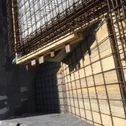 Konstrukcja zbrojeniowa i deskowanie łukowego elementu architektonicznego w trakcie budowy, widoczne bloczki betonowe i butelka wody.