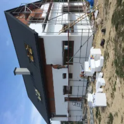 Prace przy ocieplaniu elewacji budynku jednorodzinnego. Widoczne rusztowania, pracownicy montujący izolację, białe płyty styropianowe oraz fragment dachu z czarną blachodachówką. Budynek w trakcie...