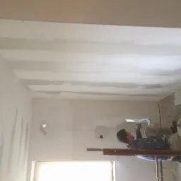 Mężczyzna na rusztowaniu, z pacą w dłoni, wyrównuje gładź na suficie w pomieszczeniu w trakcie remontu. Widoczne niedoskonałości i nierówności na ścianach, oraz wiadro z zaprawą obok.