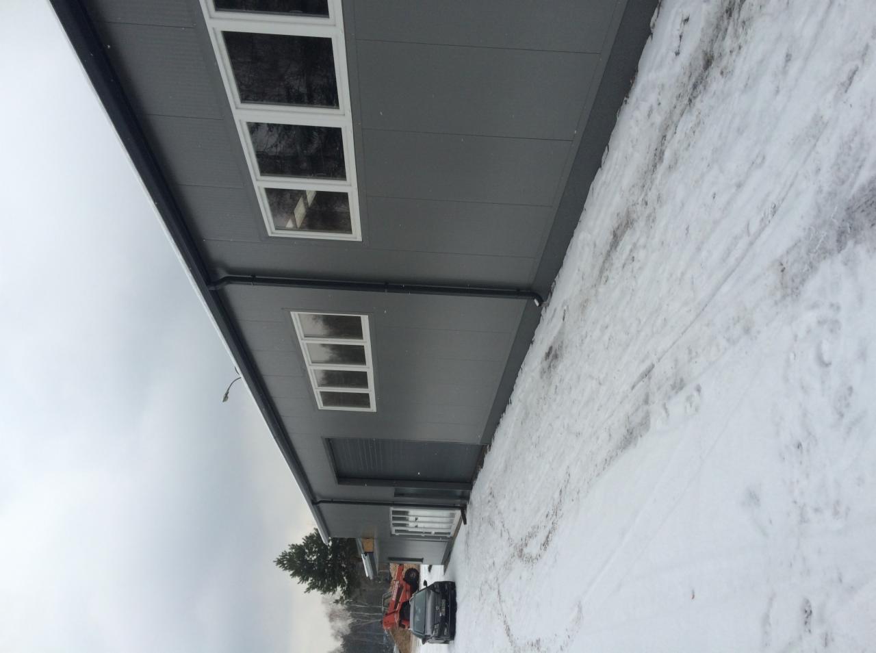 Szary budynek przemysłowy z panelowej elewacji, widoczne okna i brama garażowa, teren wokół pokryty śniegiem, pochmurne zimowe niebo.