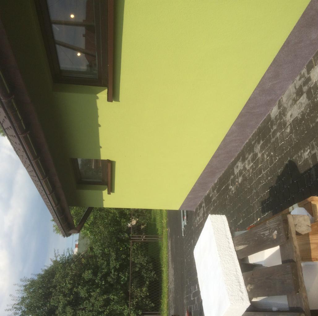 Fragment ściany domu z oknami, pomalowanej na jasnozielono, z widocznym fragmentem elewacji i kostki brukowej, obok leżą płyty styropianowe na palecie.