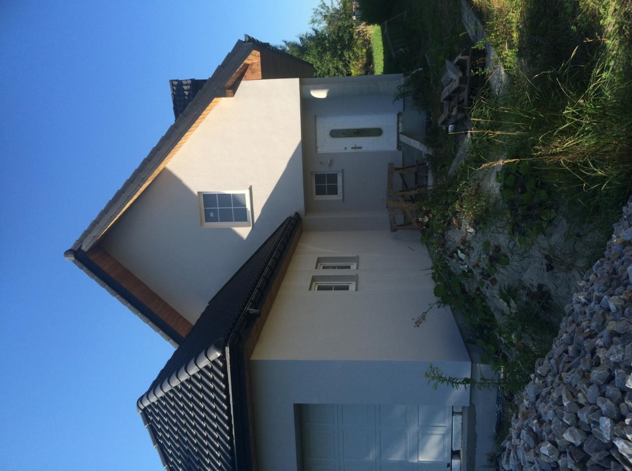 Świeżo otynkowany dom jednorodzinny z grafitową dachówką i białymi oknami, widok z poziomu gruntu na elewację frontową i boczną, z widoczną stertą kamieni i elementami roślinności.