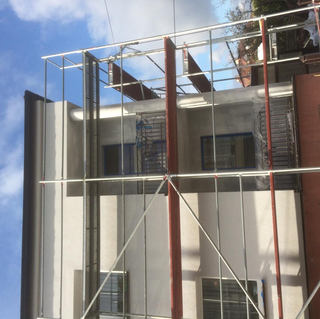 Budynek w trakcie prac elewacyjnych, widoczne rusztowanie, okna zabezpieczone folią, świeżo nałożony tynk w kolorze jasnego beżu, fragment niebieskiego nieba w tle.