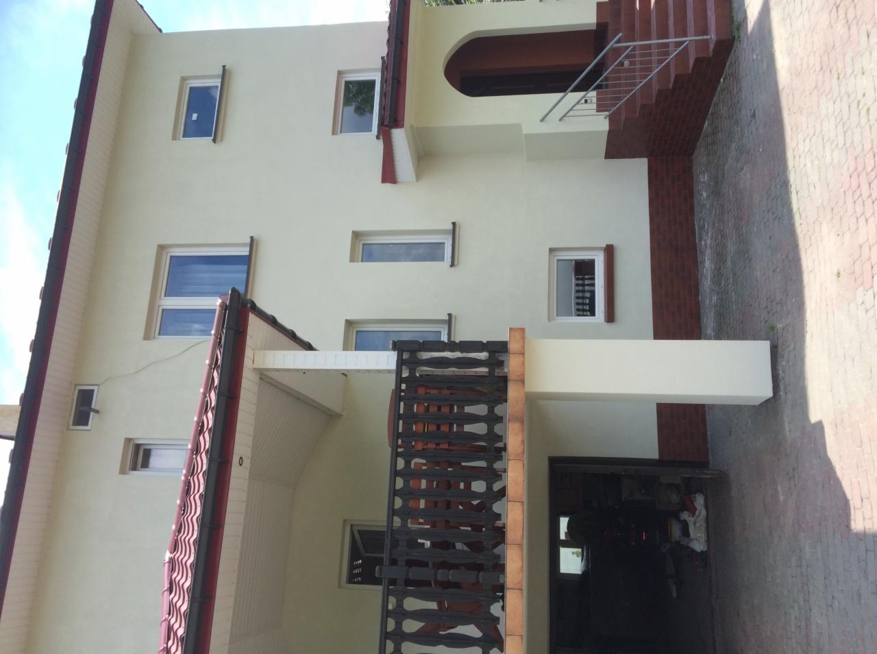 Dwupiętrowy dom z jasną elewacją, widoczne okna, balkon z ozdobną balustradą, czerwona dachówka i schody wejściowe z cegły.