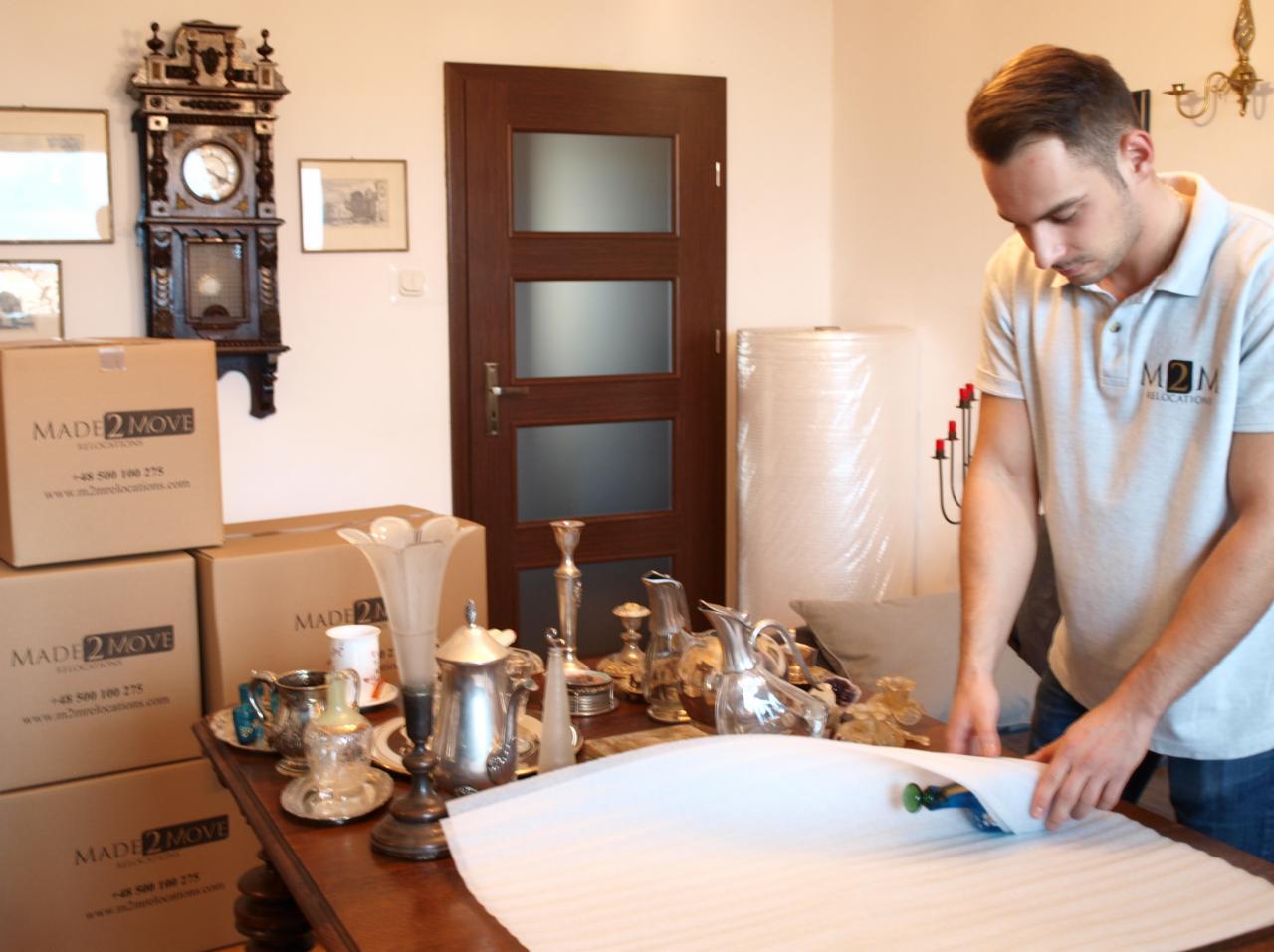 Pracownik firmy przeprowadzkowej owija delikatne przedmioty folią bąbelkową na drewnianym stole, przygotowując je do transportu; w tle widoczne kartony z logo firmy, zegar i drzwi.