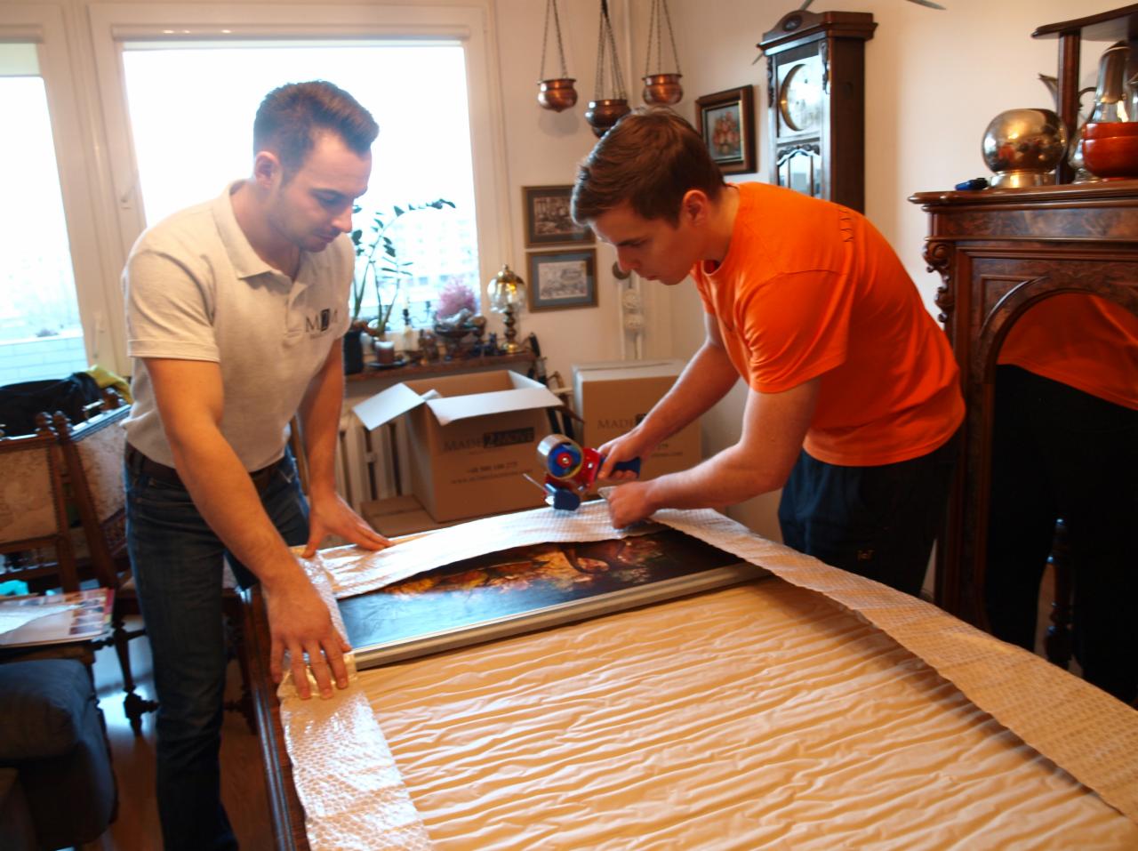 Dwóch mężczyzn zabezpiecza obraz folią bąbelkową na łóżku, przygotowując go do transportu. W tle kartony i antyczne meble.