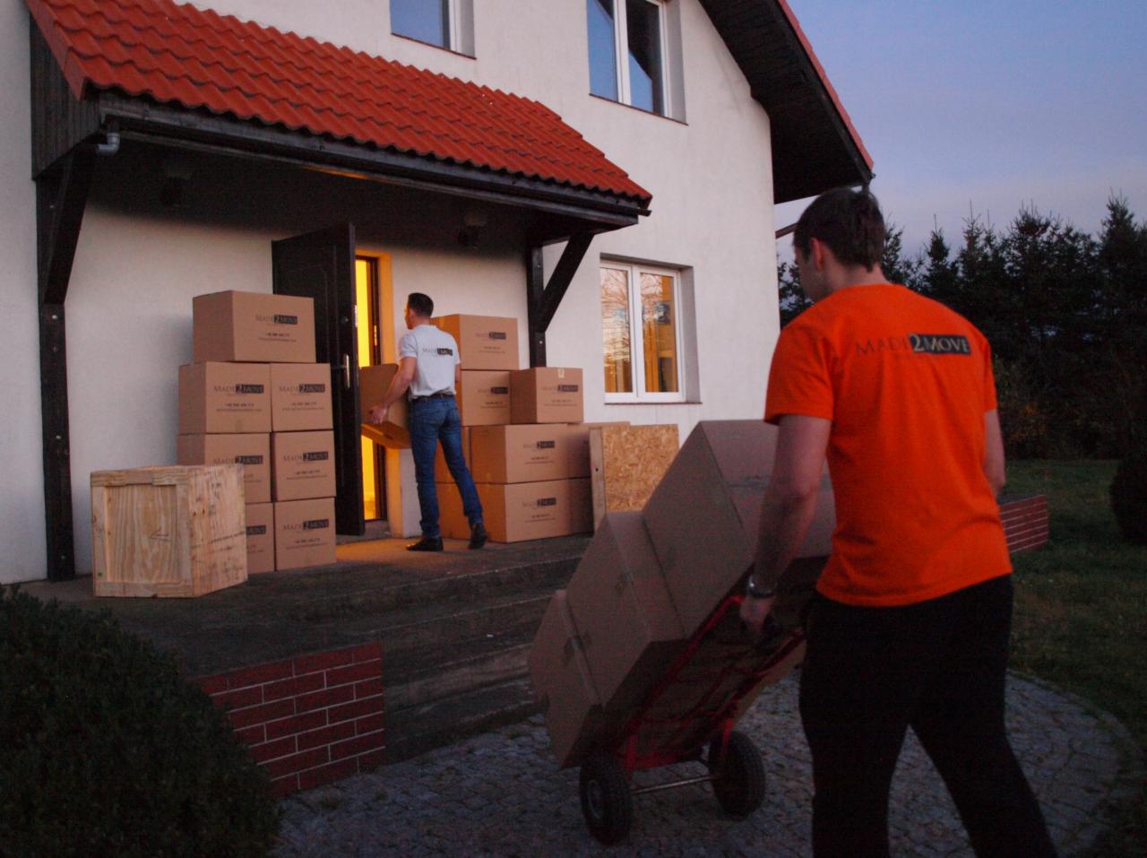 Pracownicy w trakcie przenoszenia kartonów z logo firmy na wózku transportowym; dom jednorodzinny z czerwonym dachem w tle.