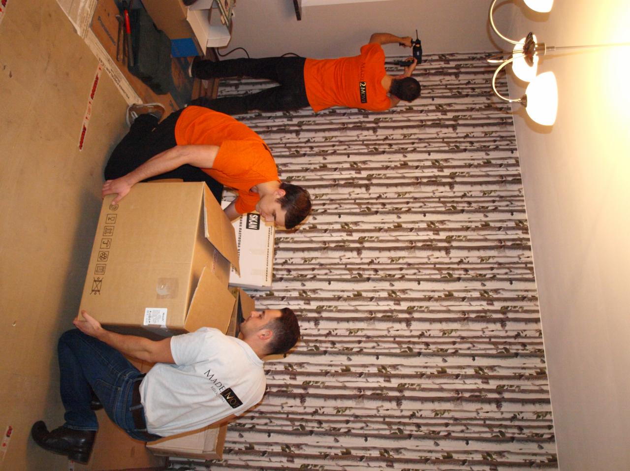 Trzech mężczyzn w firmowych koszulkach Made2Move przenosi kartony podczas przeprowadzki, jeden z nich skanuje kod kreskowy na ścianie.