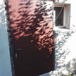 PawelSpawTech - Ciemna, metalowa brama garażowa zamontowana w surowym otworze, z widocznymi śladami zaprawy i cieniem rzucanym przez liście drzewa.