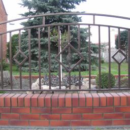 PawelSpawTech - Kuta brama ogrodzeniowa w Przygodzicach, z dekoracyjnymi rombami i łukiem, osadzona na niskim murku z czerwonej cegły, w tle zielony trawnik i drzewa.