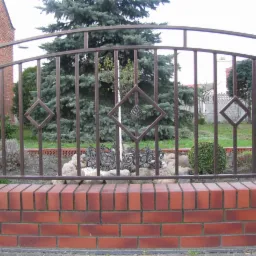 Kuta brama ogrodzeniowa w Przygodzicach, z dekoracyjnymi rombami i łukiem, osadzona na niskim murku z czerwonej cegły, w tle zielony trawnik i drzewa.