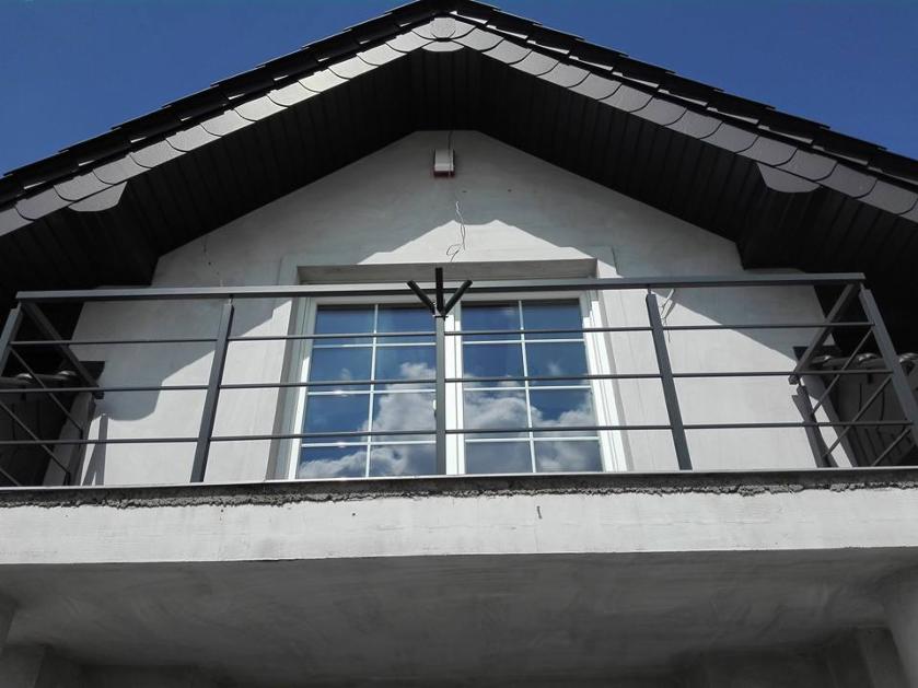 Balkon z metalową balustradą na tle białej fasady domu z ciemnym dachem, w oknach odbijają się chmury.