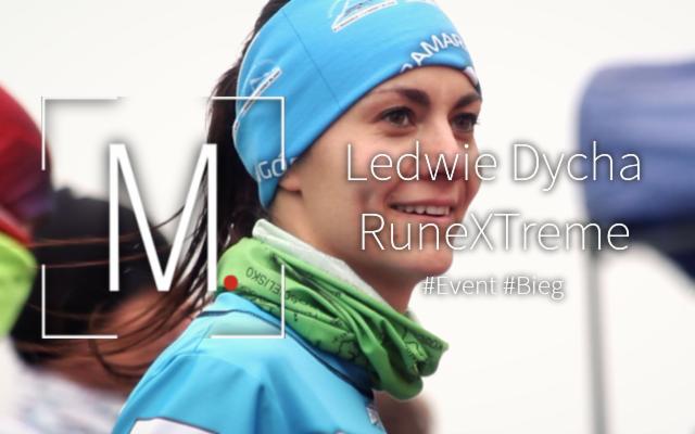 Uśmiechnięta kobieta w sportowej odzieży, z niebieską opaską na głowie i zielonym buffem na szyi, w tle rozmyty krajobraz z elementami namiotów i innych uczestników wydarzenia sportowego.