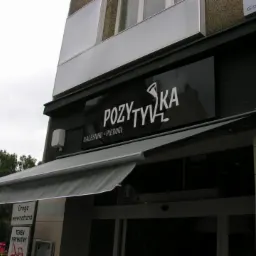 Czarny szyld restauracji 'Pozytywka' z białym napisem i logo, informujący o serwowaniu naleśników i pierogów, umieszczony nad wejściem z szarą markizą.