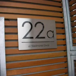 Metalowa tabliczka z numerem 22a i adresem 'ul. Nadmorski Dwór' umieszczona na drewnianej elewacji budynku.