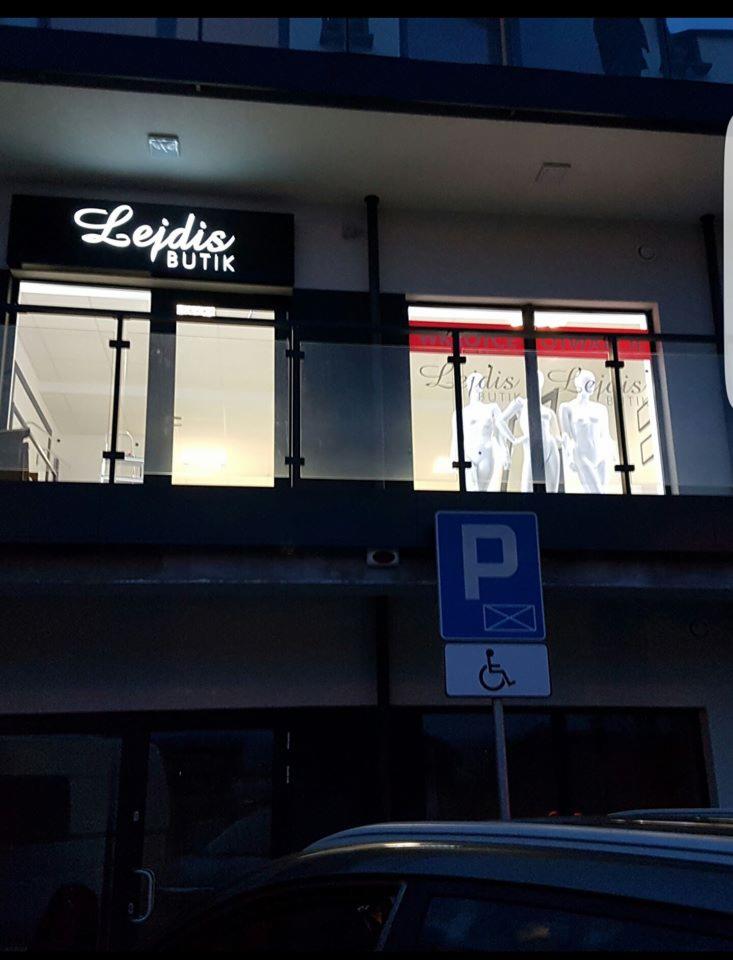 Elegancki szyld butiku 'Lejdis' z podświetlanym logo nad witryną, na tle nowoczesnego budynku z miejscem parkingowym dla niepełnosprawnych.