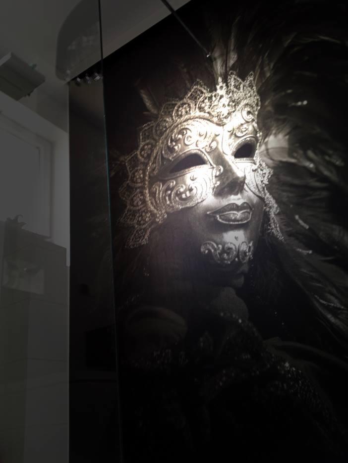 Czarno-biała fotografia dekoracyjnej weneckiej maski z piórami, umieszczona na szklanym panelu.