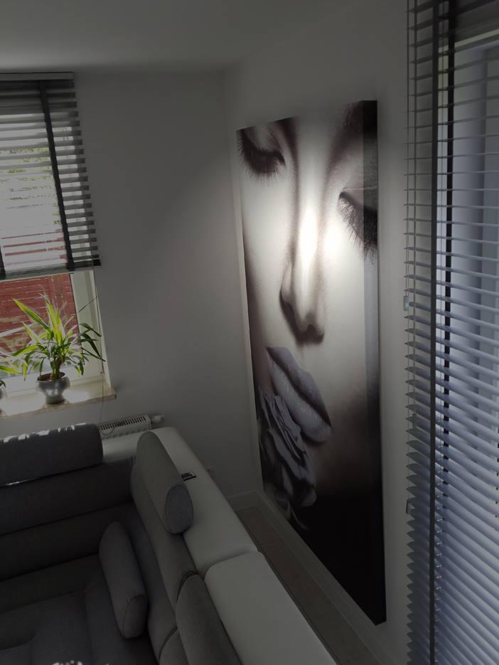 Wnętrze salonu w Rumi z dużym, pionowym, czarno-białym portretem kobiety na ścianie, widocznym za narożną sofą i obok okna z żaluzjami.