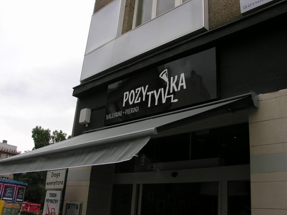 Czarny szyld restauracji 'Pozytywka' z białym napisem i logo, informujący o serwowaniu naleśników i pierogów, umieszczony nad wejściem z szarą markizą.