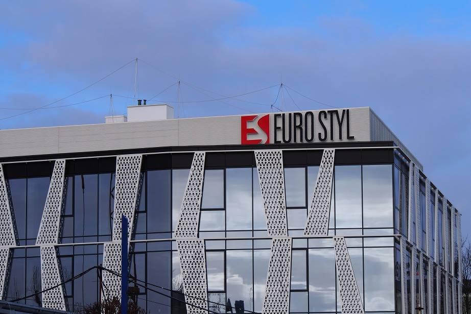 Budynek biurowy z nowoczesną fasadą, logo firmy Euro Styl na dachu, widoczne elementy dekoracyjne z ażurowym wzorem na elewacji, niebieskie niebo w tle.