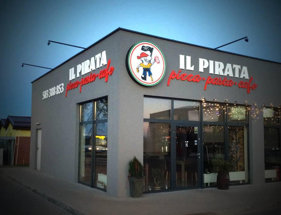 Budynek pizzerii 'Il Pirata' z dużym okrągłym szyldem z piratem, czerwonym napisem 'pizza pasta cafe' i numerem telefonu na szarej elewacji.