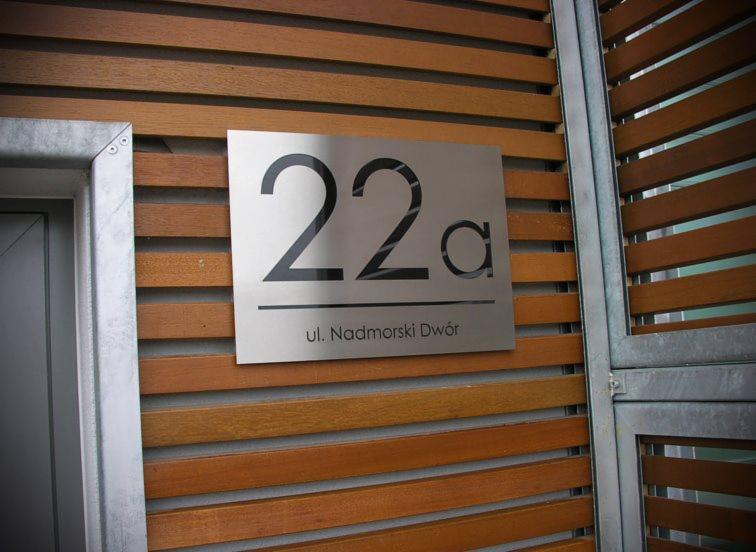 Metalowa tabliczka z numerem 22a i adresem 'ul. Nadmorski Dwór' umieszczona na drewnianej elewacji budynku.