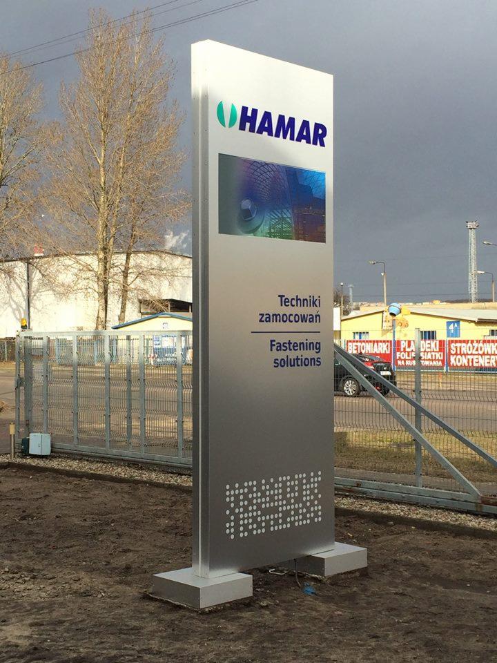 Pylon reklamowy firmy Hamar z logo, zdjęciem produktu i hasłem Techniki Zamocowań, umieszczony przed siedzibą firmy na tle pochmurnego nieba.