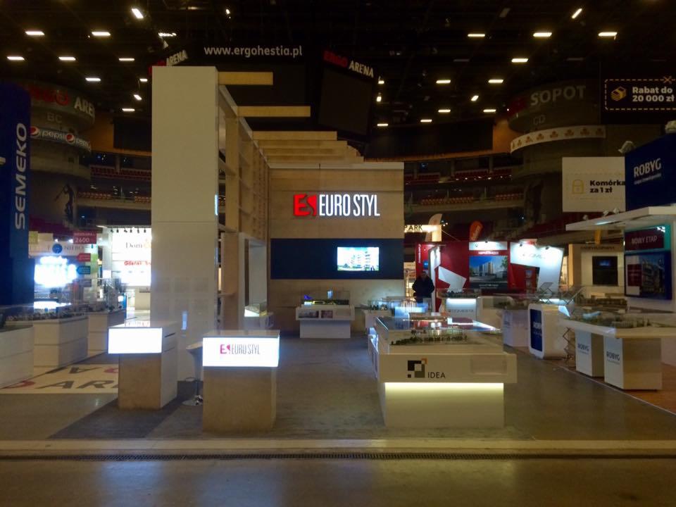 Stoisko targowe z podświetlanym logo 'EURO STYL' i modelami architektonicznymi, widoczne inne stoiska i trybuny hali.