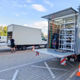 Dwa busy transportowe na parkingu z kostki brukowej. Jeden bus z otwartą rampą załadunkową i metalową konstrukcją wewnątrz. Pracownicy w czarnych koszulkach i spodenkach przygotowują się...