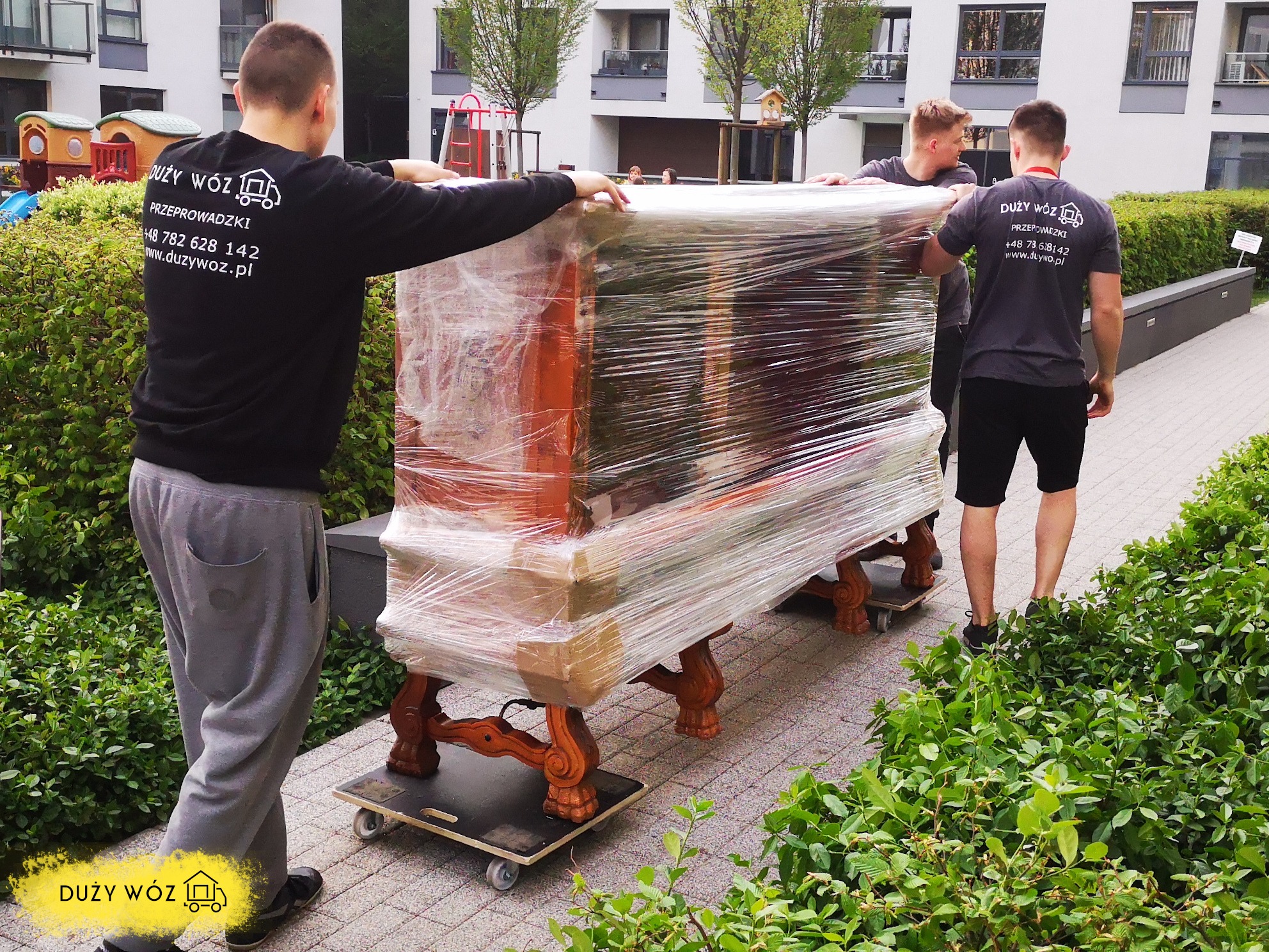 Trzech pracowników firmy przeprowadzkowej w Warszawie transportuje owinięty folią antyczny stół na wózku po chodniku przed budynkiem mieszkalnym.