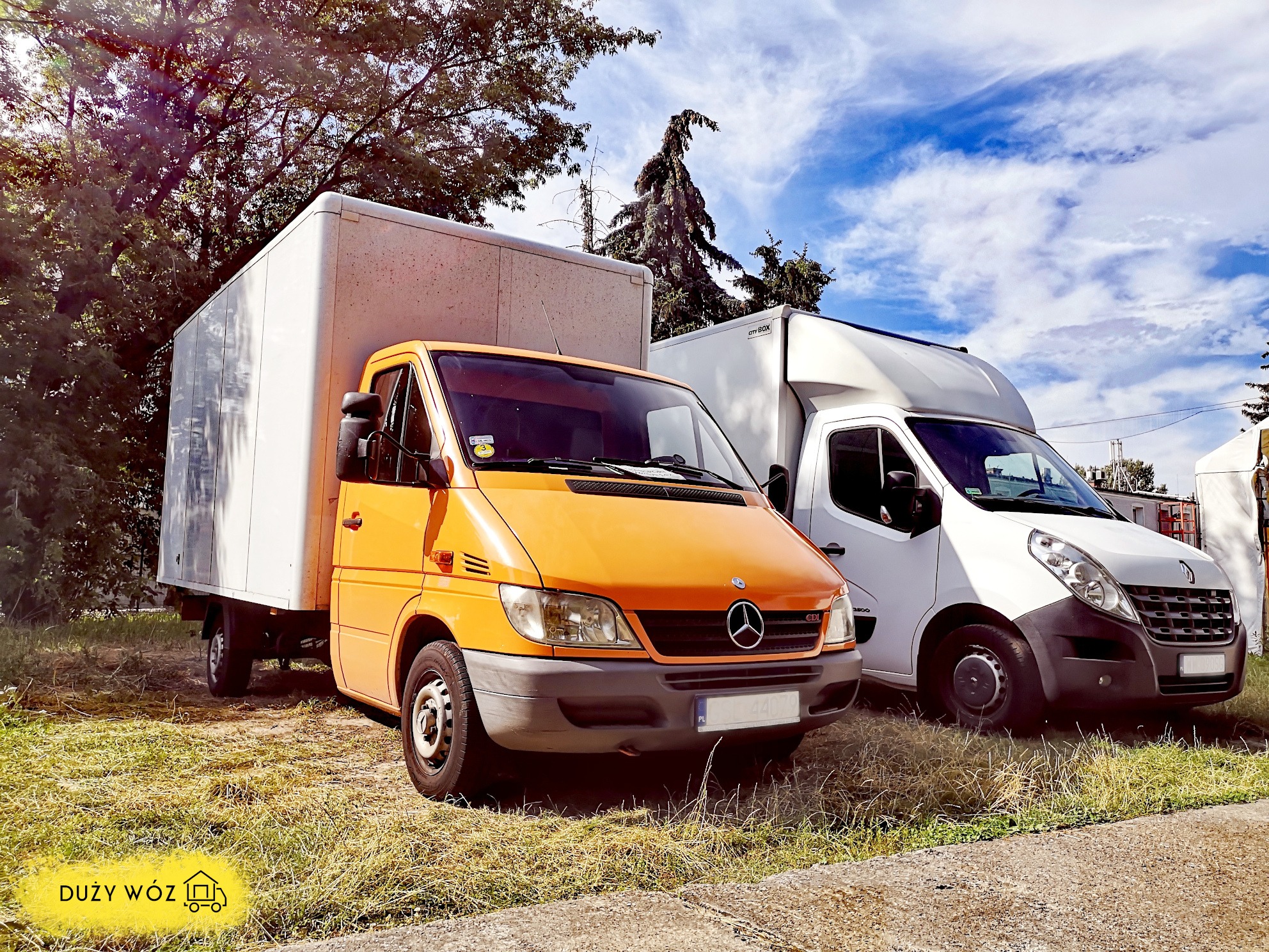 Dwa busy dostawcze: pomarańczowy Mercedes-Benz Sprinter z zabudową kontenerową i biały Renault Master, zaparkowane na trawiastym terenie pod drzewami, z widocznym logo firmy transportowej w lewym...