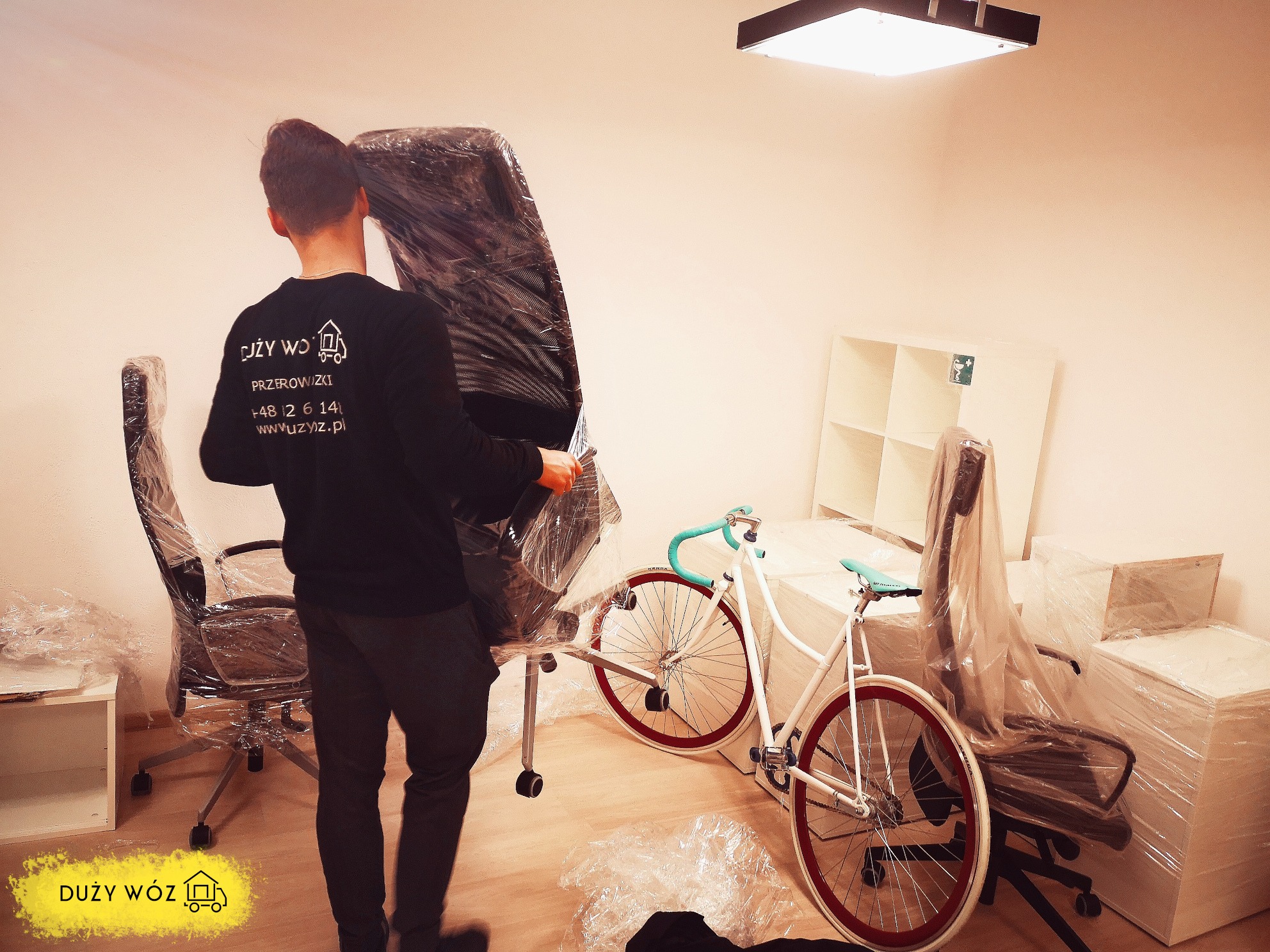 Pracownik firmy przeprowadzkowej w czarnej bluzie z logo na plecach przenosi zapakowane w folię krzesło biurowe; w tle zapakowane meble i rower.