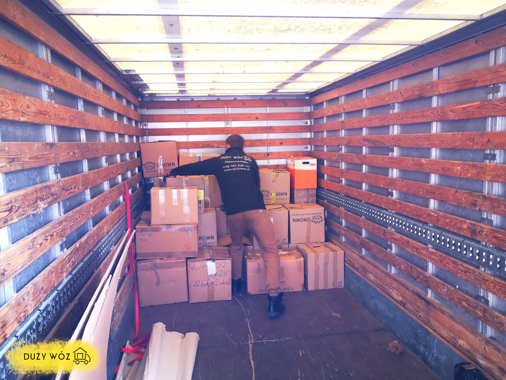 Wnętrze furgonetki wypełnione kartonami podczas załadunku, pracownik w czarnej bluzie z logo firmy układa pudełka, widoczne drewniane zabezpieczenia ścian pojazdu.